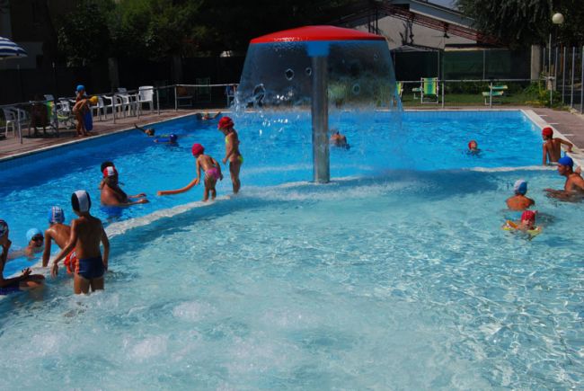 Bambini che giocano in piscina con una fontana centrale.