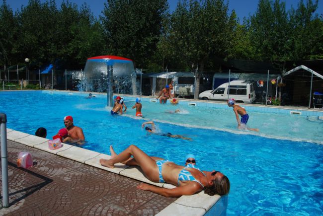 Persone nuotano in piscina, una donna prende il sole sul bordo.