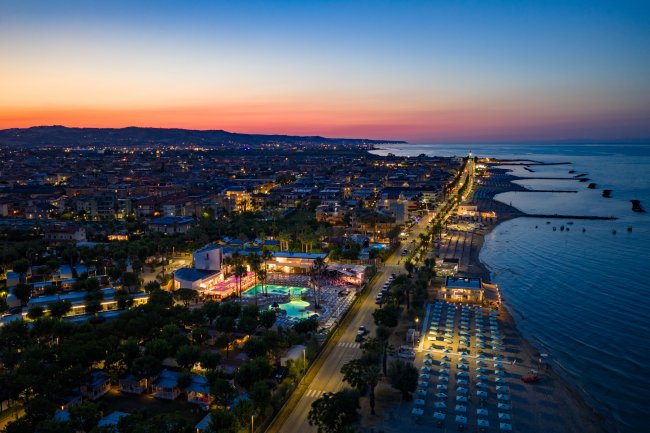 Spiaggia al tramonto con file di ombrelloni e luci accese lungo la costa.