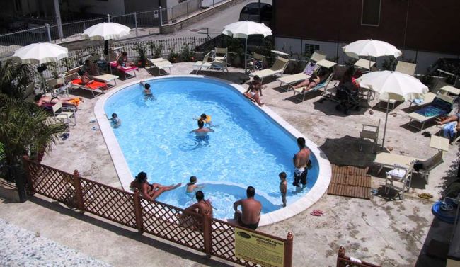 Piscina all'aperto con persone che nuotano e prendono il sole sotto ombrelloni bianchi.