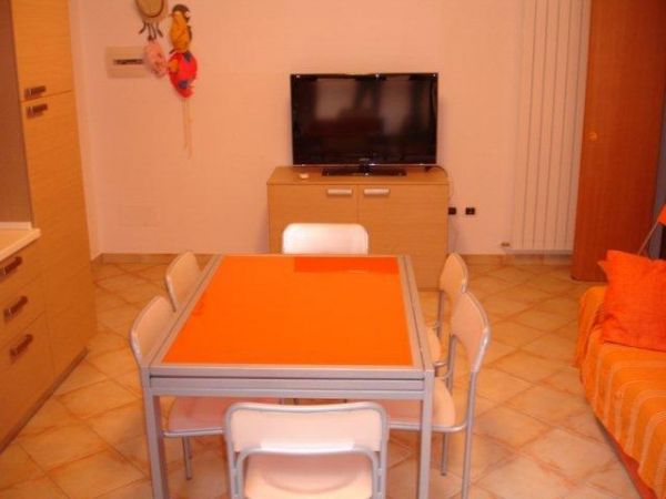 Soggiorno con tavolo arancione, sedie bianche e TV su mobile di legno chiaro.