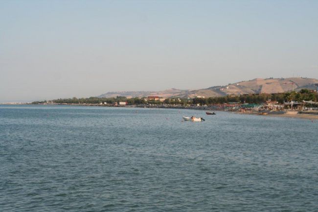 Spiaggia sabbiosa e colline in lontananza, con mare calmo e barche.